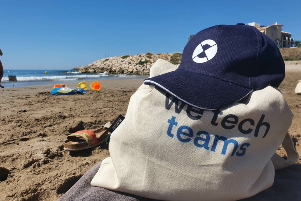 Una gorra corporativa de CrossPoint apoyada sobre una bolsa de tela con el lema We tech teams en la arena de la playa
