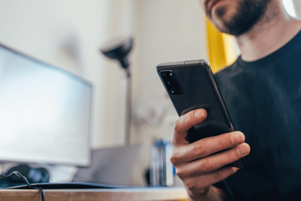 Un chico revisando su tel&eacute;fono m&oacute;vil, con el ordenador de fondo